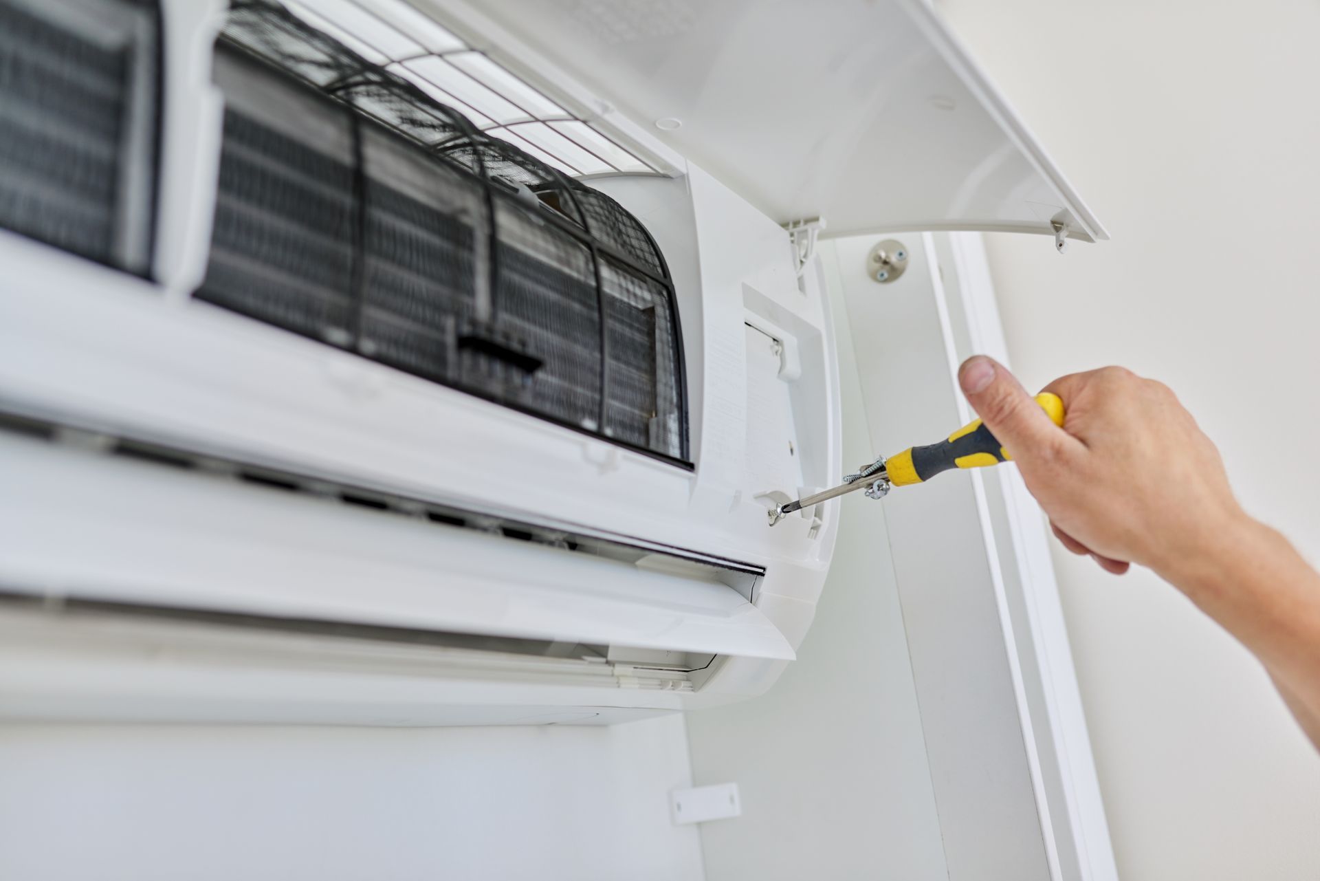 A person uses a yellow-handled screwdriver to repair the casing of a wall-mounted air conditioning unit.