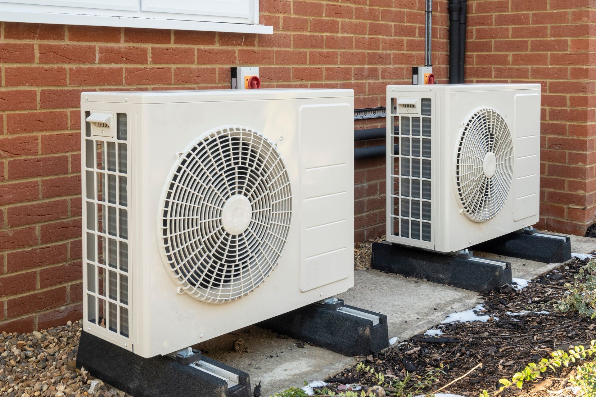 Two beige, industrial outdoor heat pump units sit on black mounting blocks against a red brick wall.