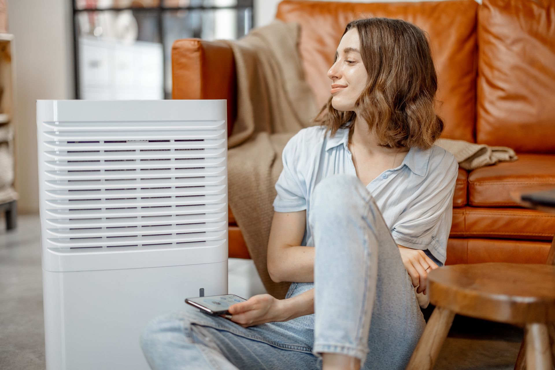 A person sits on a living room floor holding a remote, next to a white air purifier, in front of a brown leather couch.
