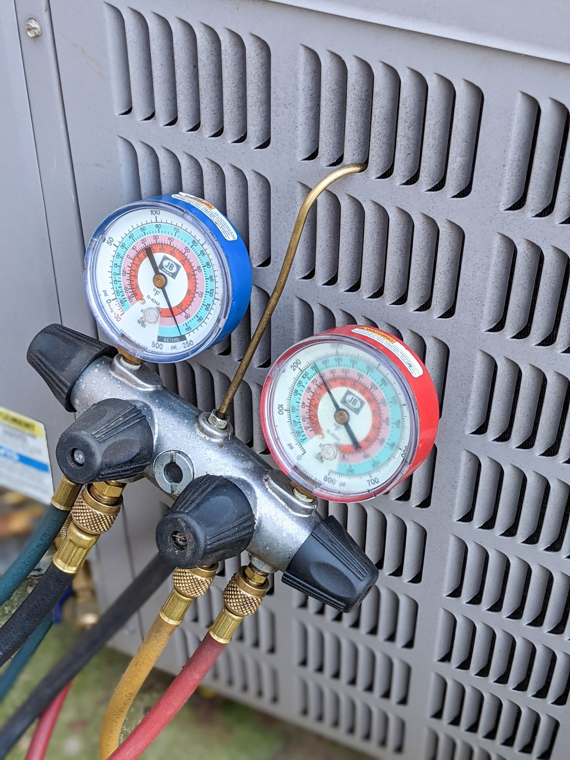 HVAC manifold gauge set with red and blue pressure dials connected to the side of an outdoor air conditioning unit.