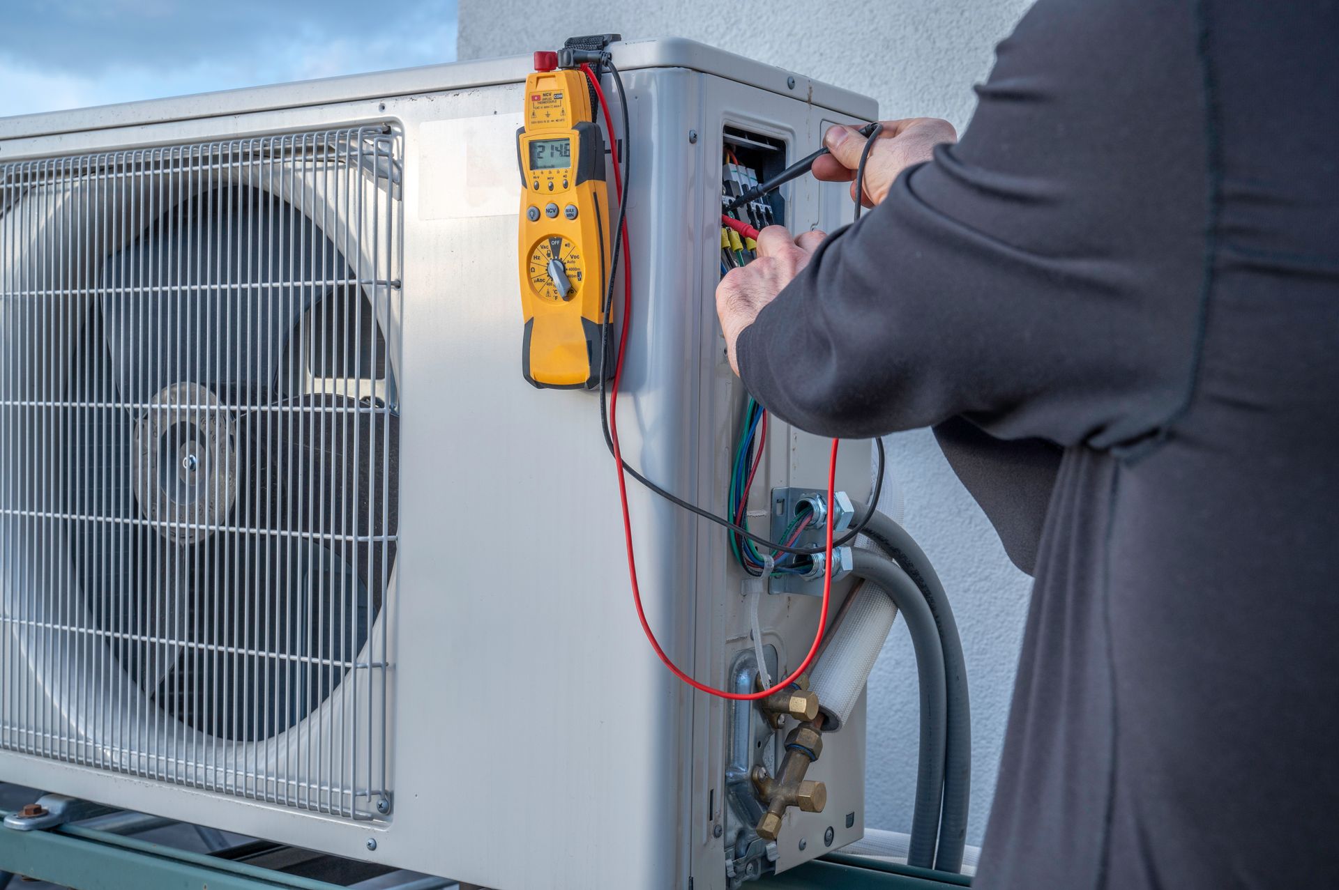 A technician uses a yellow multimeter to test electrical wiring inside an outdoor HVAC condenser unit.