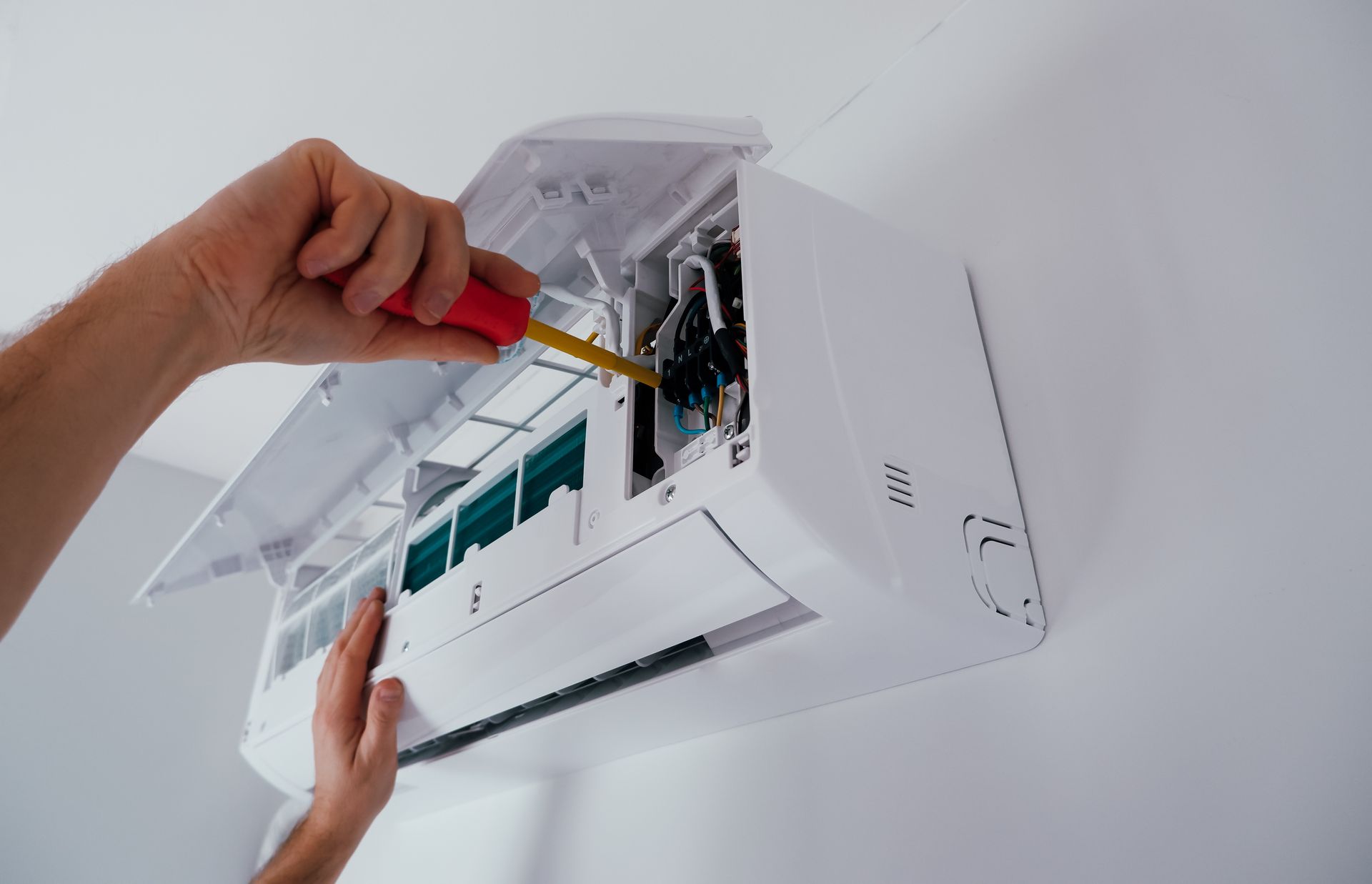 A person uses a red-handled screwdriver to repair the internal electrical components of a wall-mounted air conditioner.