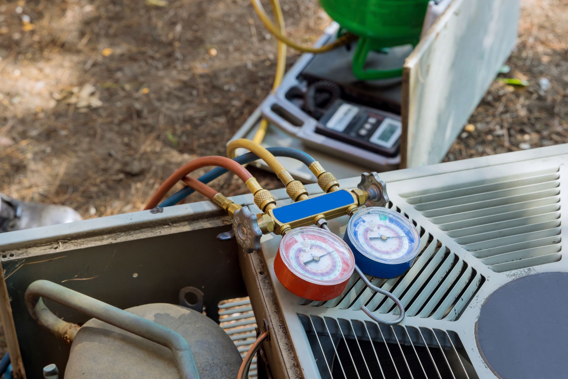 HVAC technician tools, including a manifold gauge set with red and blue dials, connected to an outdoor air conditioner.