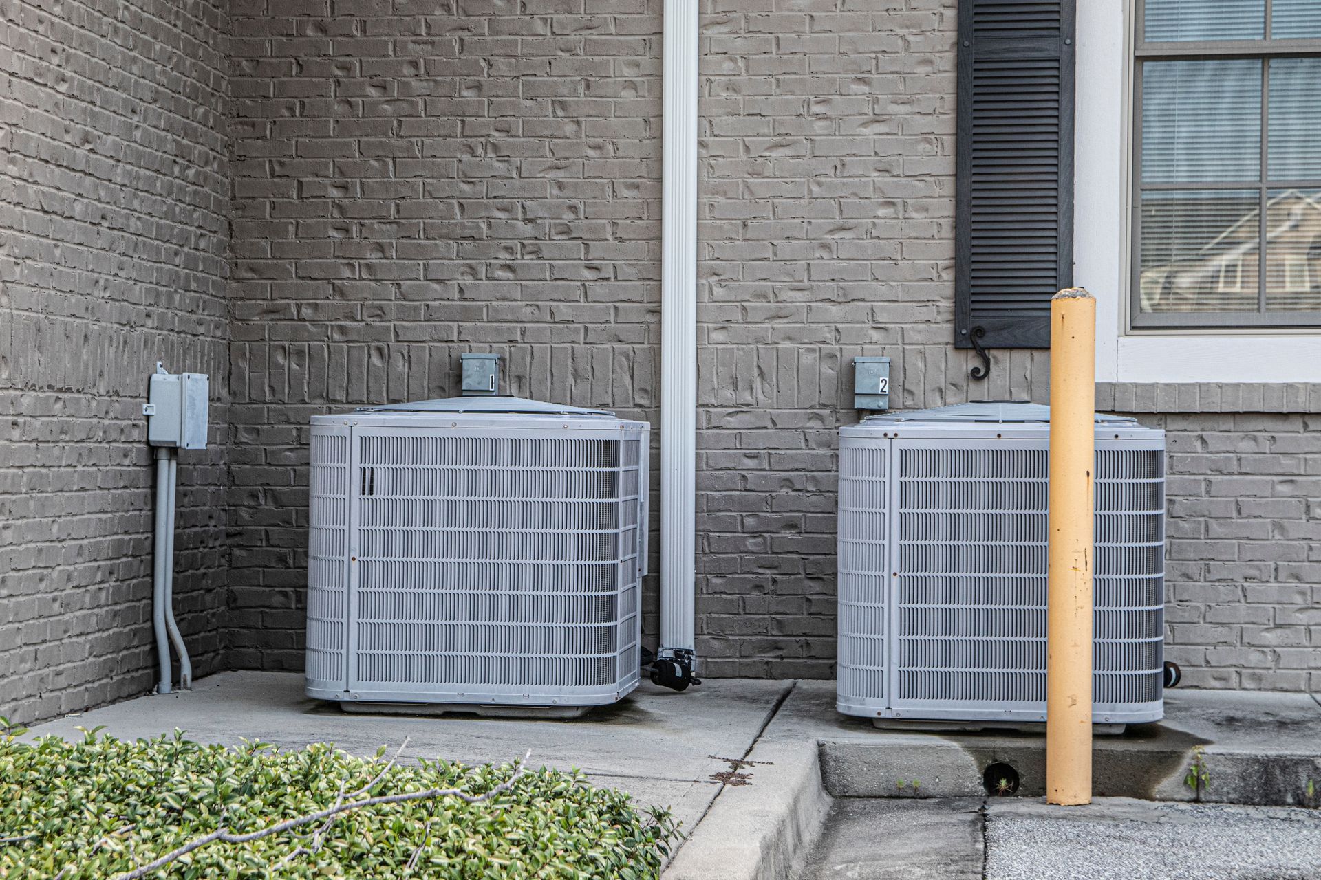 Two outdoor HVAC condenser units sit on a concrete pad against a gray brick wall, with a yellow bollard on the right.