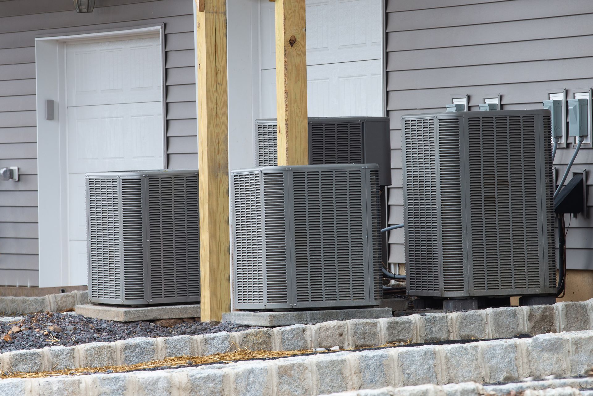 Three gray HVAC condenser units sit on concrete pads outside a gray-sided building with wooden support posts.