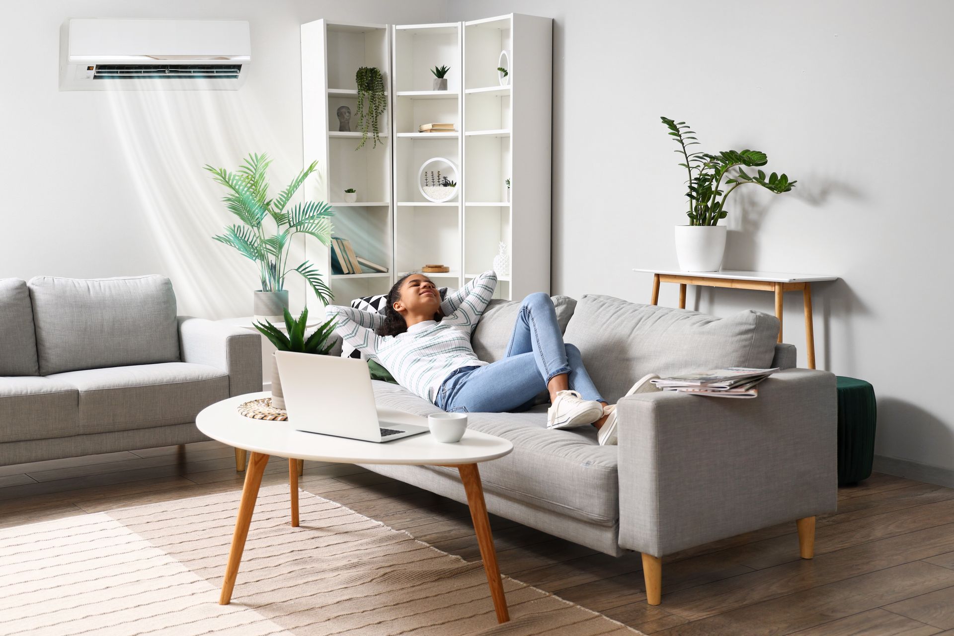 A person rests on a gray sofa in a brightly lit, modern living room while an air conditioner blows air downward.
