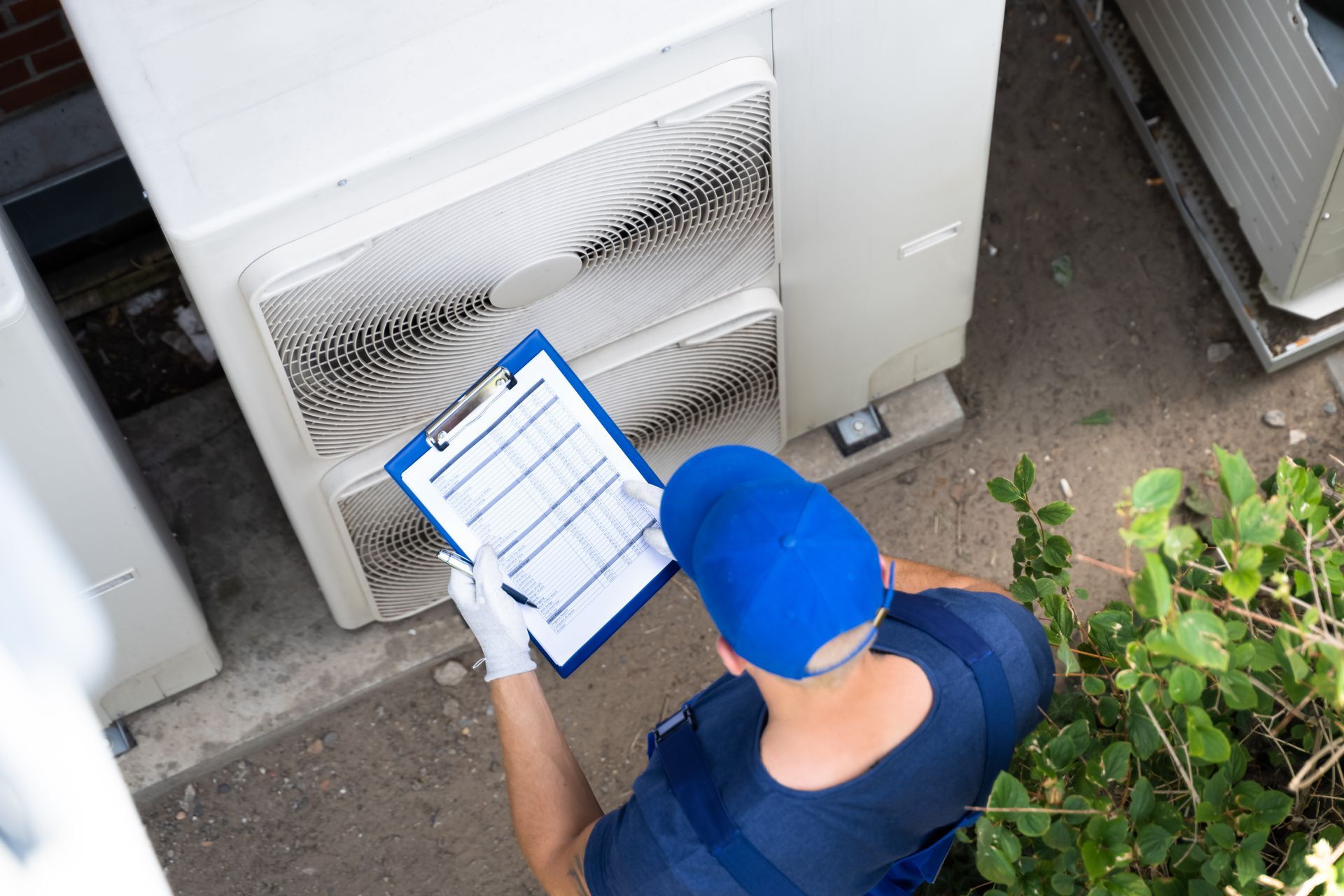 A person wearing a blue cap and work uniform stands outdoors, inspecting an air conditioning unit while writing on a clipboard.