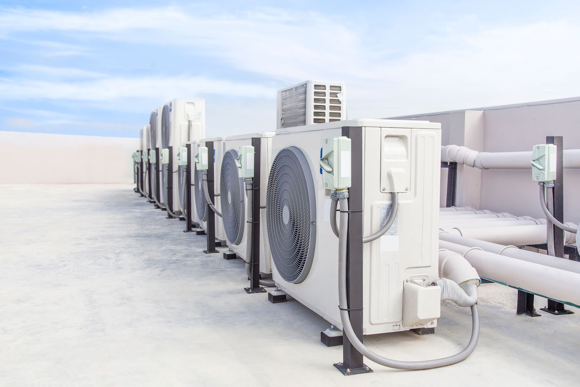 A row of white outdoor HVAC air conditioning units installed on a flat, light-colored rooftop against a blue sky.
