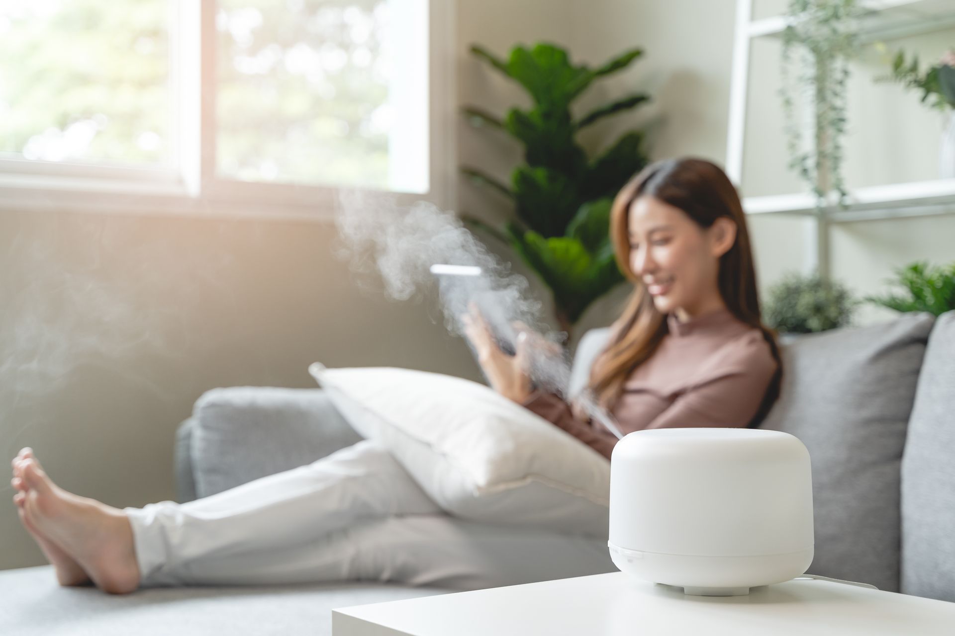 A person sits on a sofa, smiling while looking at a tablet with a white aromatherapy diffuser in the foreground.