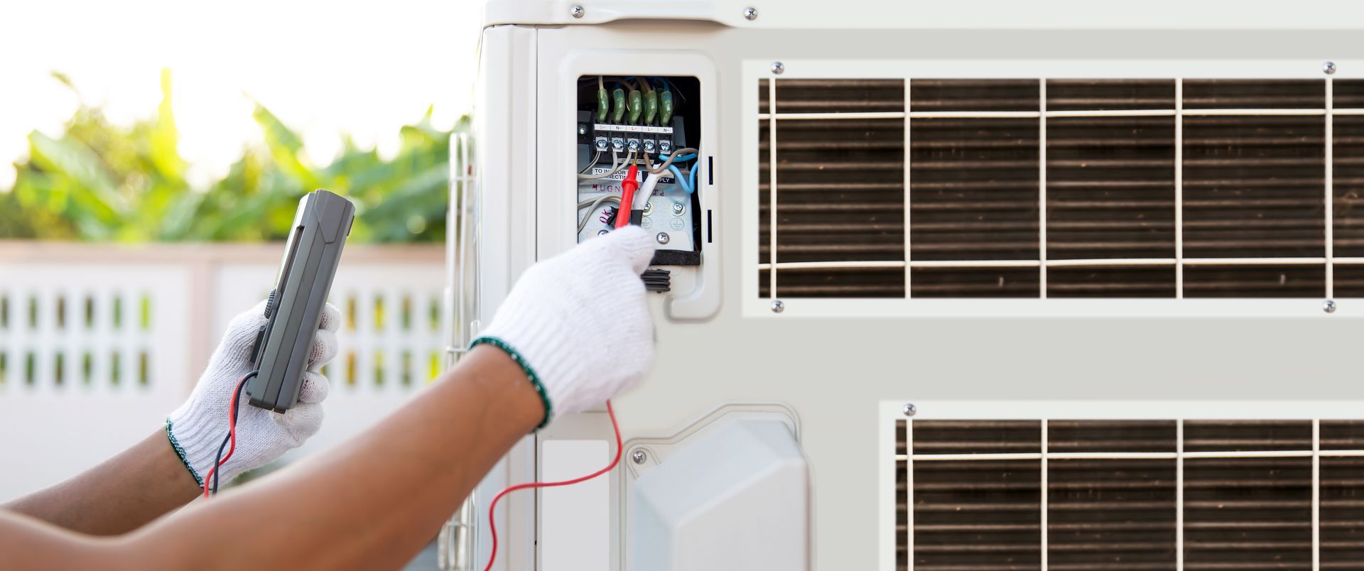 A technician in white gloves uses a multimeter to test the electrical wiring of an outdoor air conditioning unit.