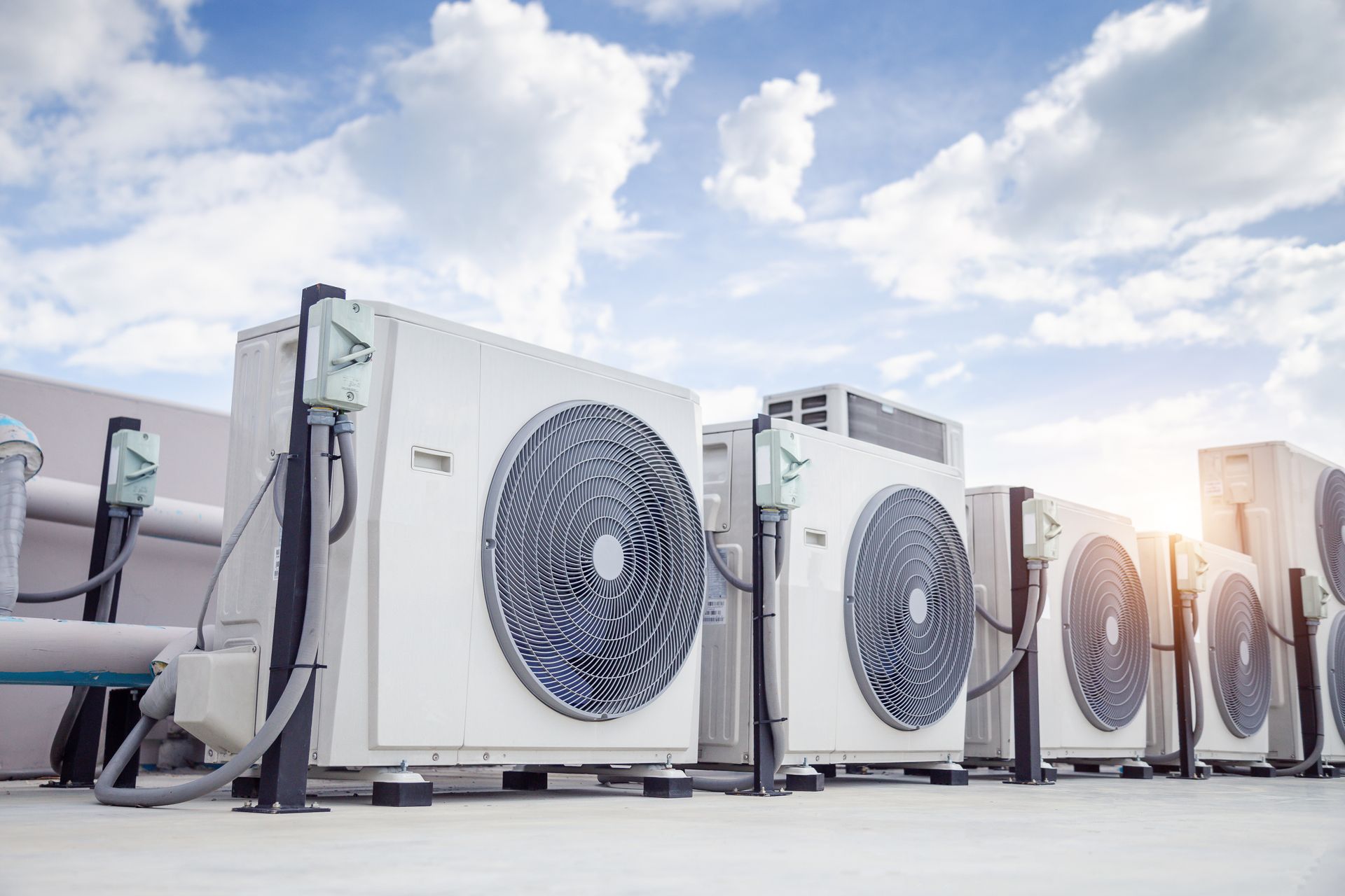 A row of white industrial air conditioning units positioned outdoors against a cloudy blue sky.