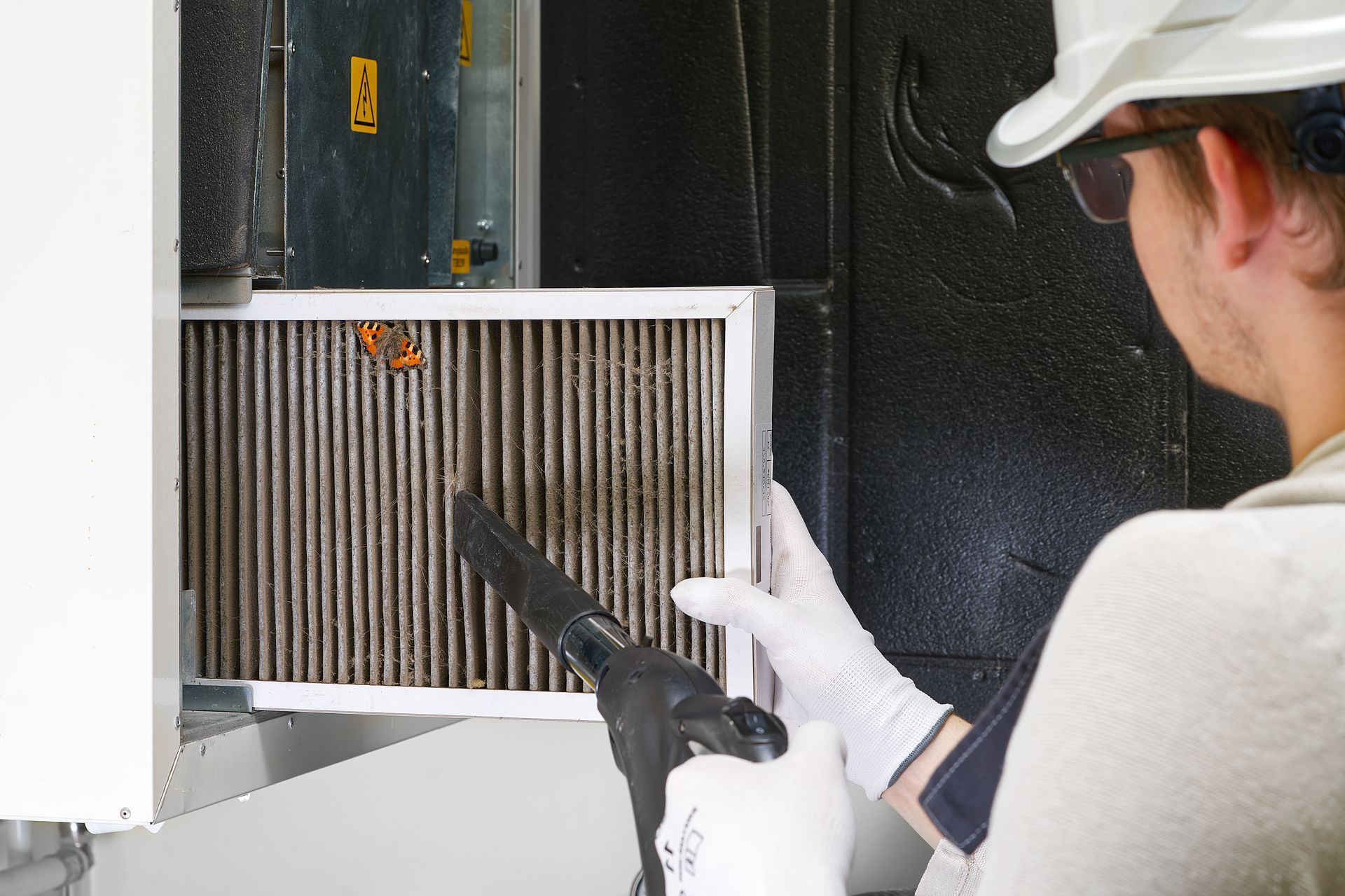 A technician wearing safety glasses, a hard hat, and gloves uses a compressed air nozzle to clean a dirty HVAC filter.