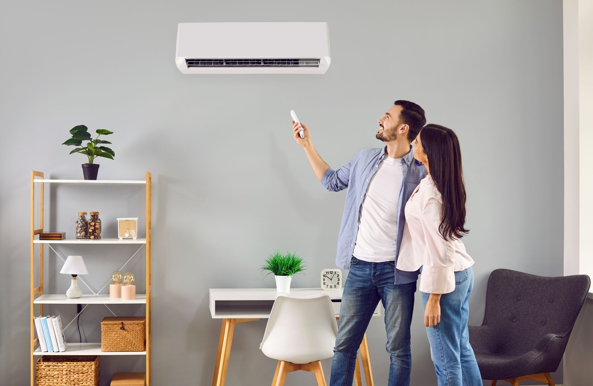 A couple stands in a living room, pointing a remote control at a wall-mounted air conditioner.