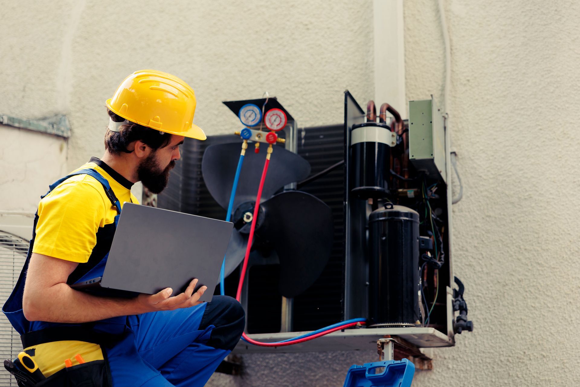A technician in a yellow hard hat and uniform checks an outdoor HVAC unit using a laptop and manifold gauges.