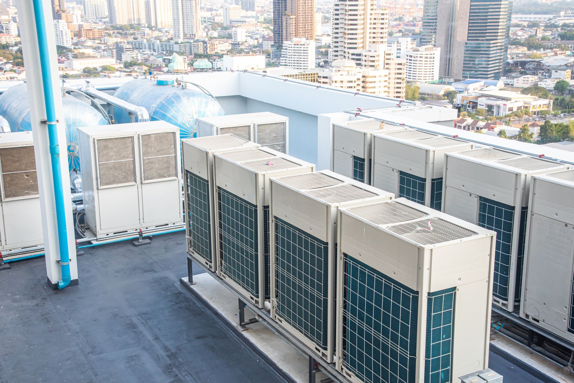 A row of large, beige industrial HVAC units on a rooftop with a city skyline in the background.