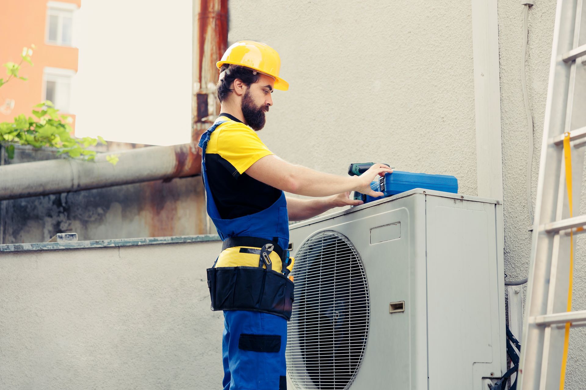 A technician in a hard hat and tool belt servicing an outdoor HVAC condenser unit next to a ladder
