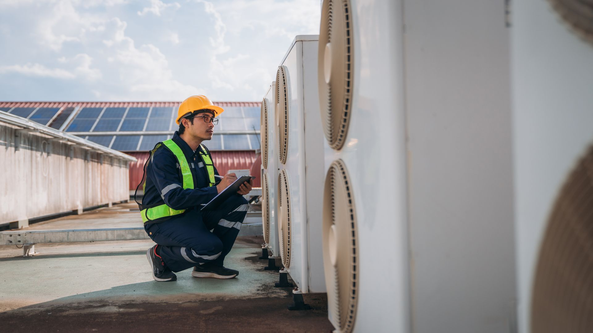 A technician in a hard hat and high-visibility vest inspects outdoor HVAC units on a rooftop.
