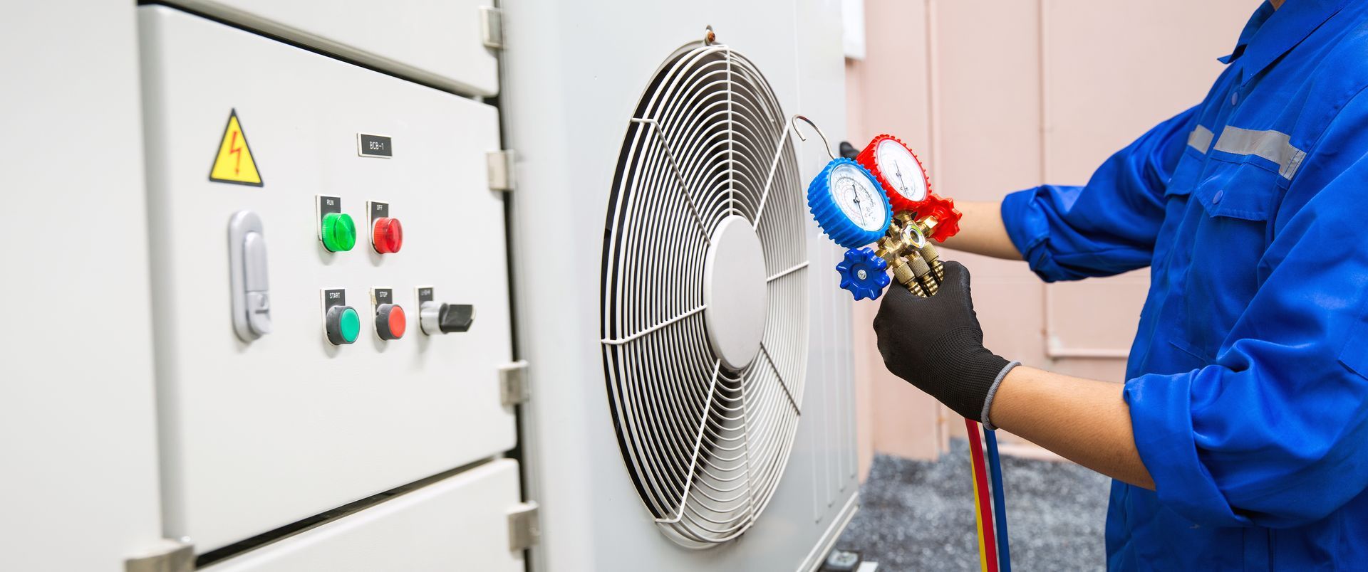 A technician in a blue uniform checks an outdoor HVAC unit with a manifold gauge set.