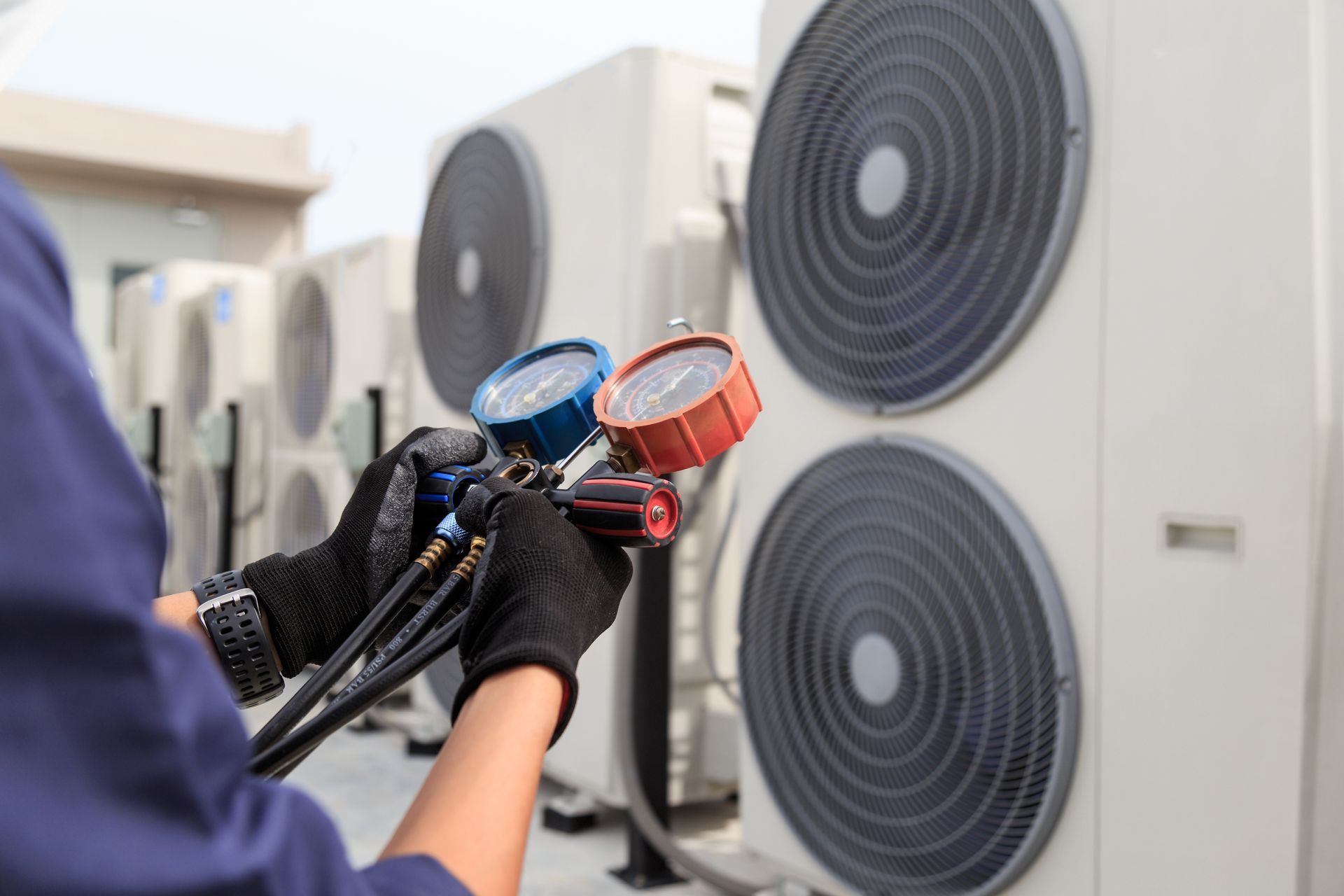A technician in work gloves holds a manifold gauge set while servicing outdoor air conditioning units.