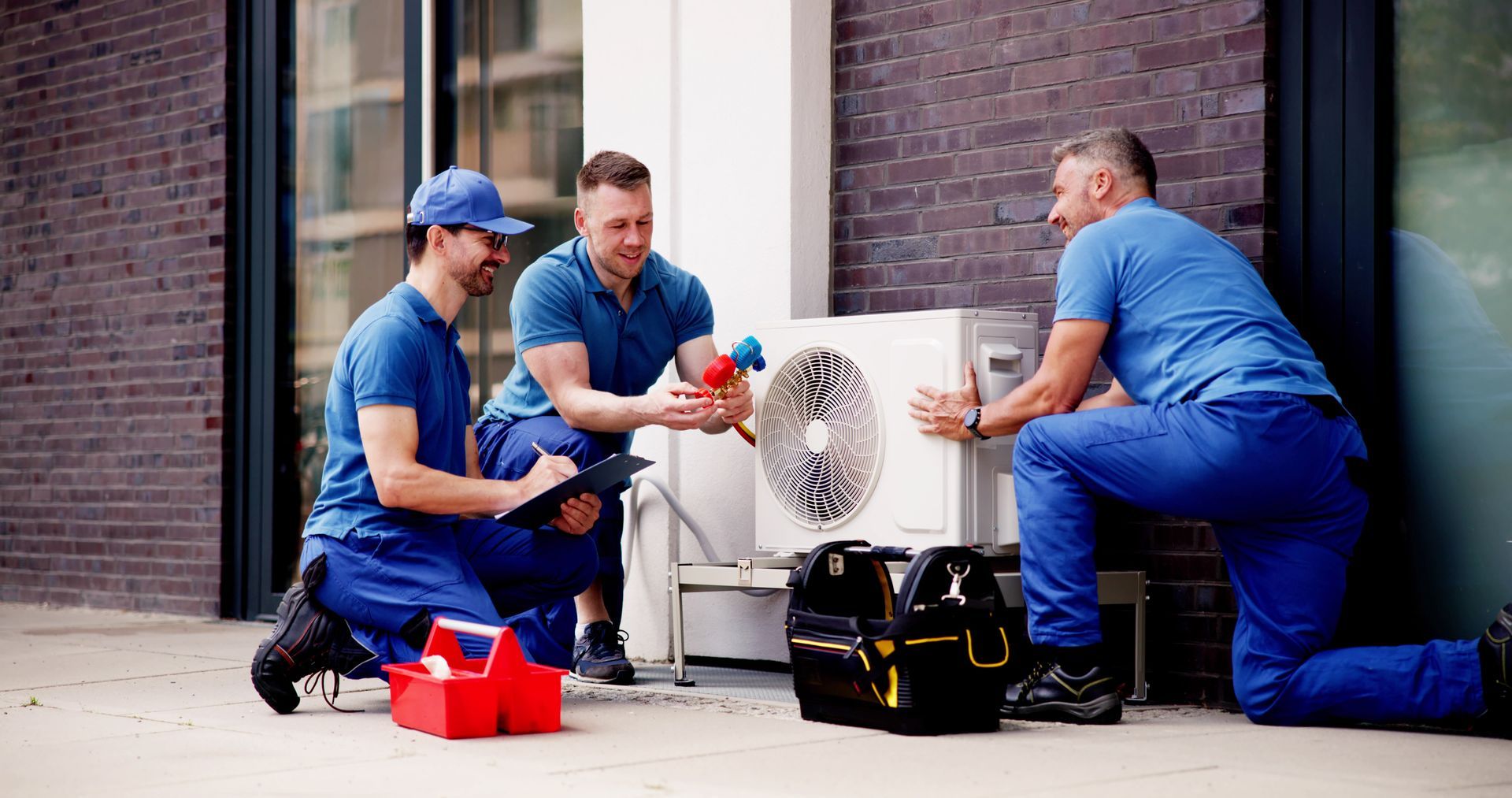 Three technicians in blue uniforms work together to repair an outdoor HVAC unit against a brick wall.