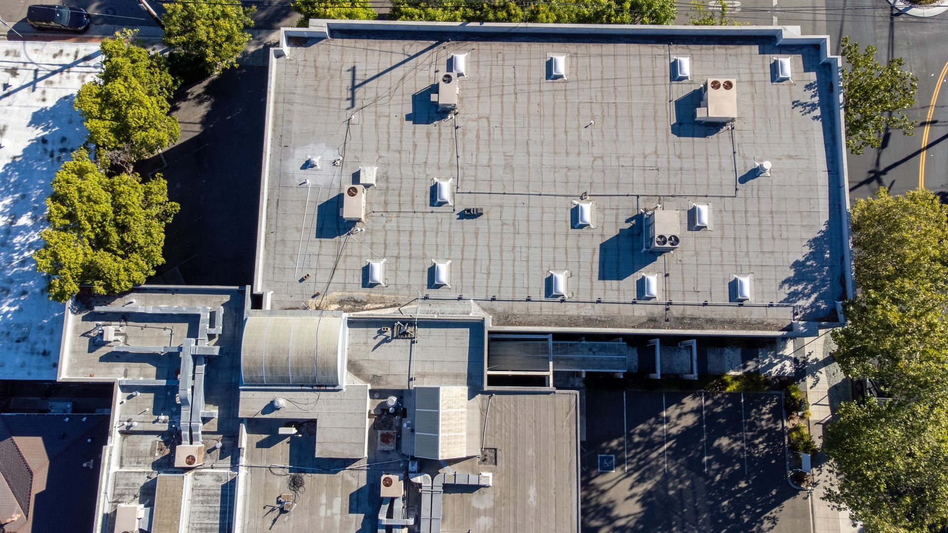 Aerial view of a flat commercial roof with multiple vents and HVAC units, surrounded by trees and a parking lot.
