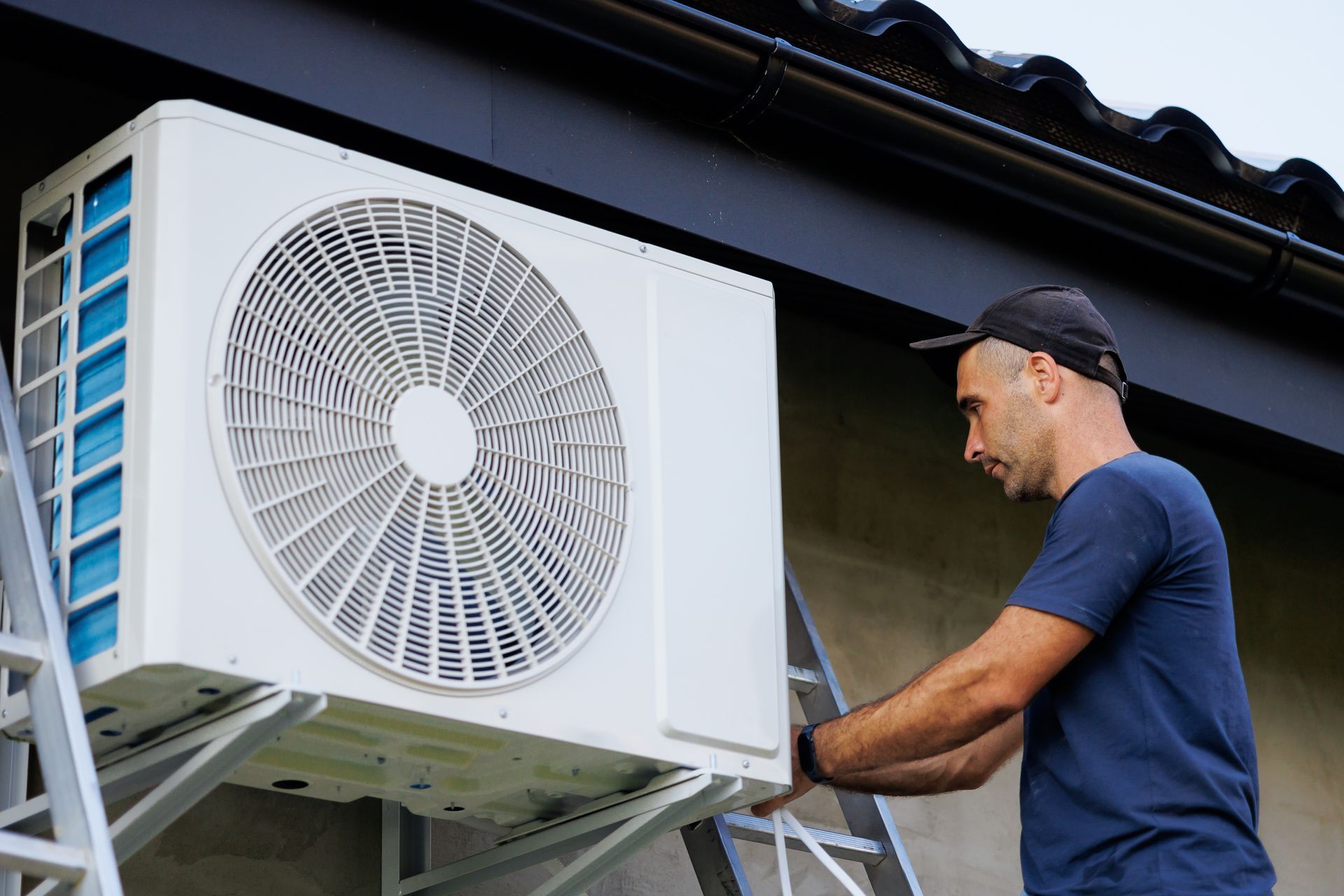 A technician in a blue shirt installs a white outdoor air conditioning unit on a building wall using a ladder.