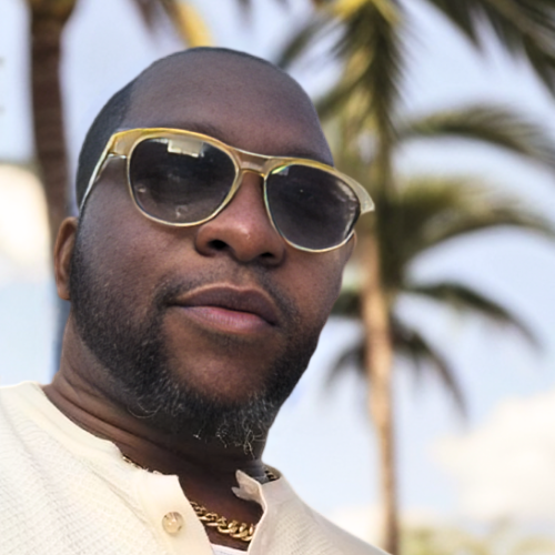 A person wearing sunglasses and a light-colored shirt, posed outdoors with palm trees in the background.