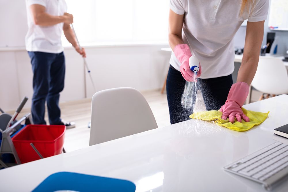 Two people cleaning an office. One sprays a desk with a cleaner while wearing pink gloves and wiping with a yellow cloth. Another is mopping.