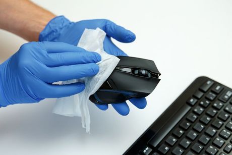Person in blue gloves wipes a computer mouse with a sanitizing wipe near a keyboard.