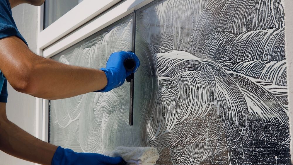 Person in blue gloves using a squeegee to clean a soapy window.