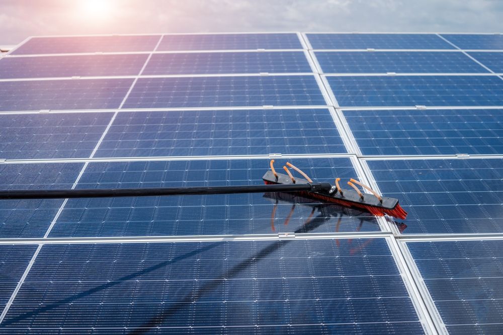 A solar panel being cleaned with a long-handled brush against a partly cloudy sky.