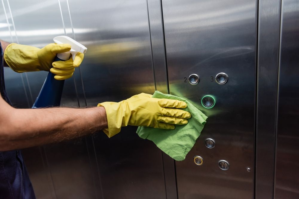 Person wearing yellow gloves spraying and wiping down elevator buttons with a green cloth.