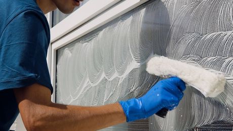Person in blue gloves cleaning a window with soapy water using a squeegee.