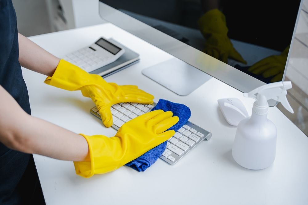 Person wearing yellow gloves cleans a computer keyboard with a blue cloth, white desk.