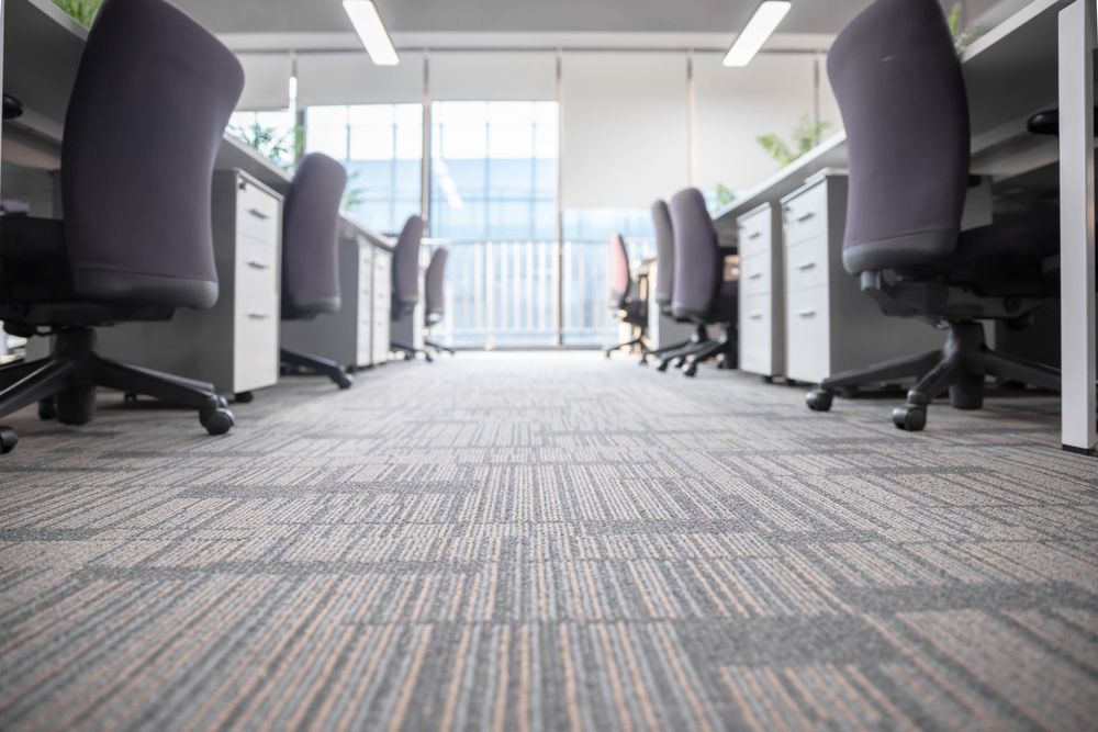 Office cubicles with gray carpet, chairs, and desks, beside large windows.