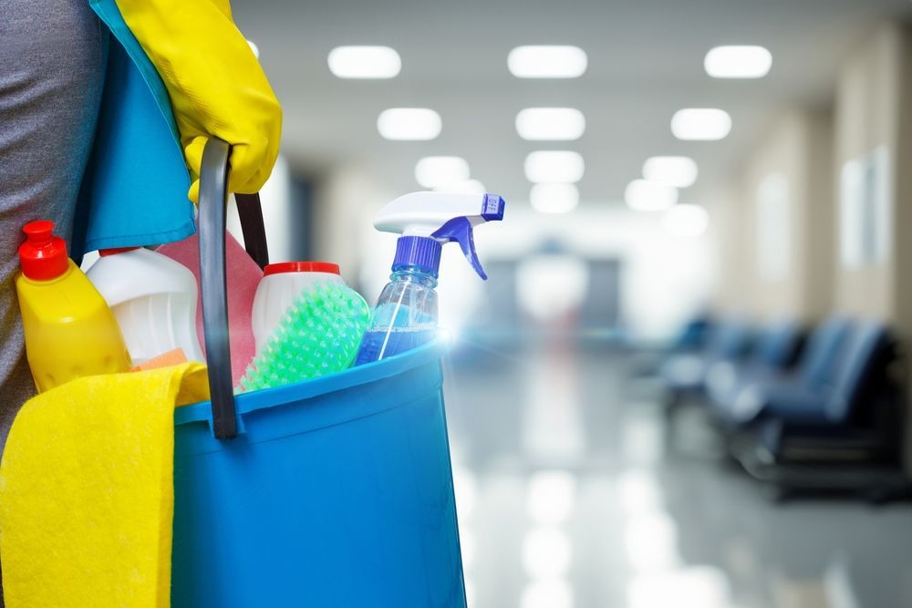 Person holding a cleaning bucket with supplies in a hallway; yellow gloves, blue bucket, blurry background.