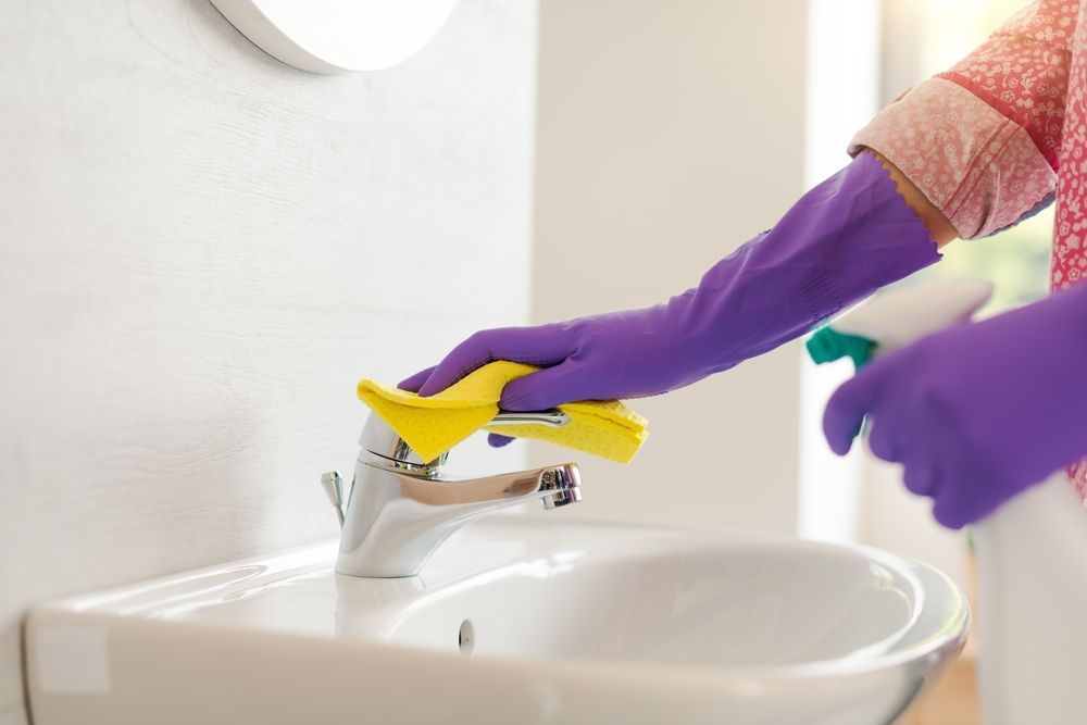 Person wearing purple gloves cleans a chrome faucet in a white bathroom using a yellow cloth and spray bottle.