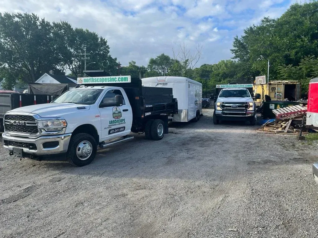 A white Ram pickup truck and a work trailer parked on a gravel lot, with a second truck and equipment in the background.