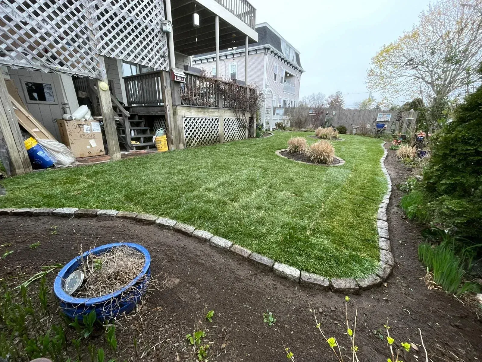 A backyard lawn with stone-edged flower beds and a blue planter in the foreground, next to a multi-story house.