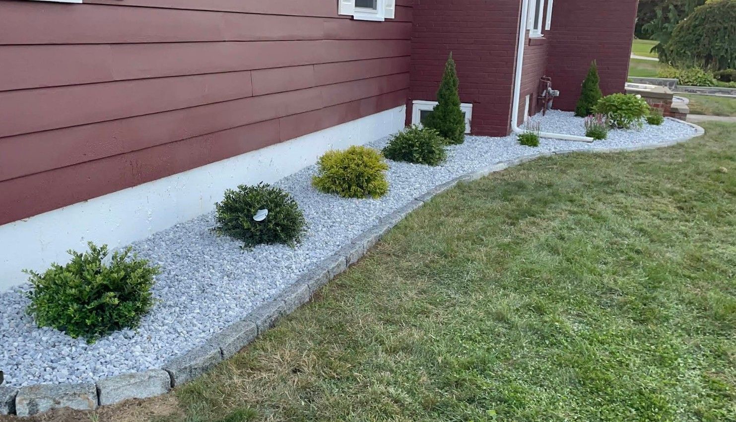 A landscaped garden bed with grey stone mulch, small green shrubs, and a stone border next to a red-sided house.