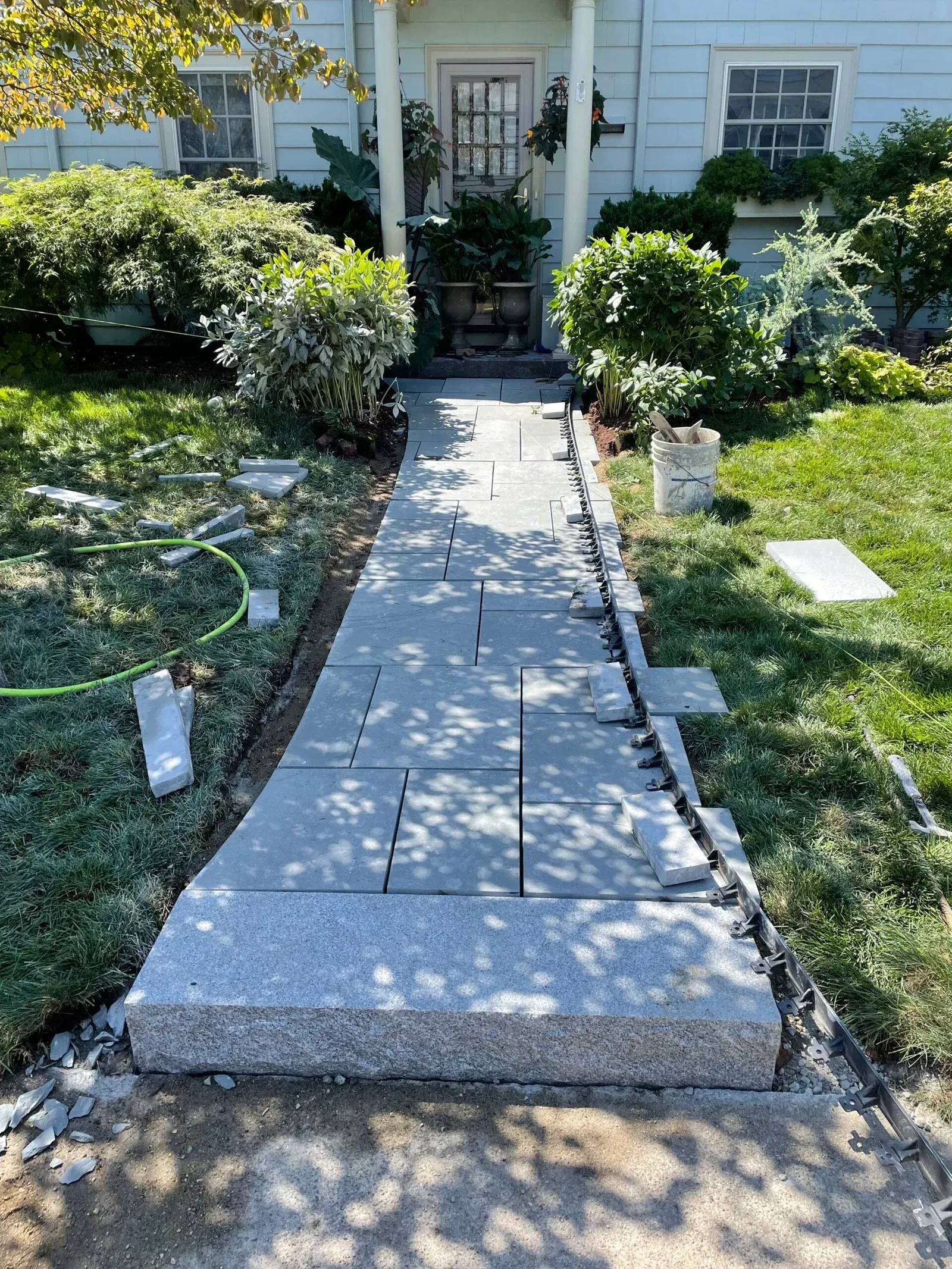 A straight, stone-paver walkway leading to the front door of a light-blue house, flanked by green bushes and lawn.