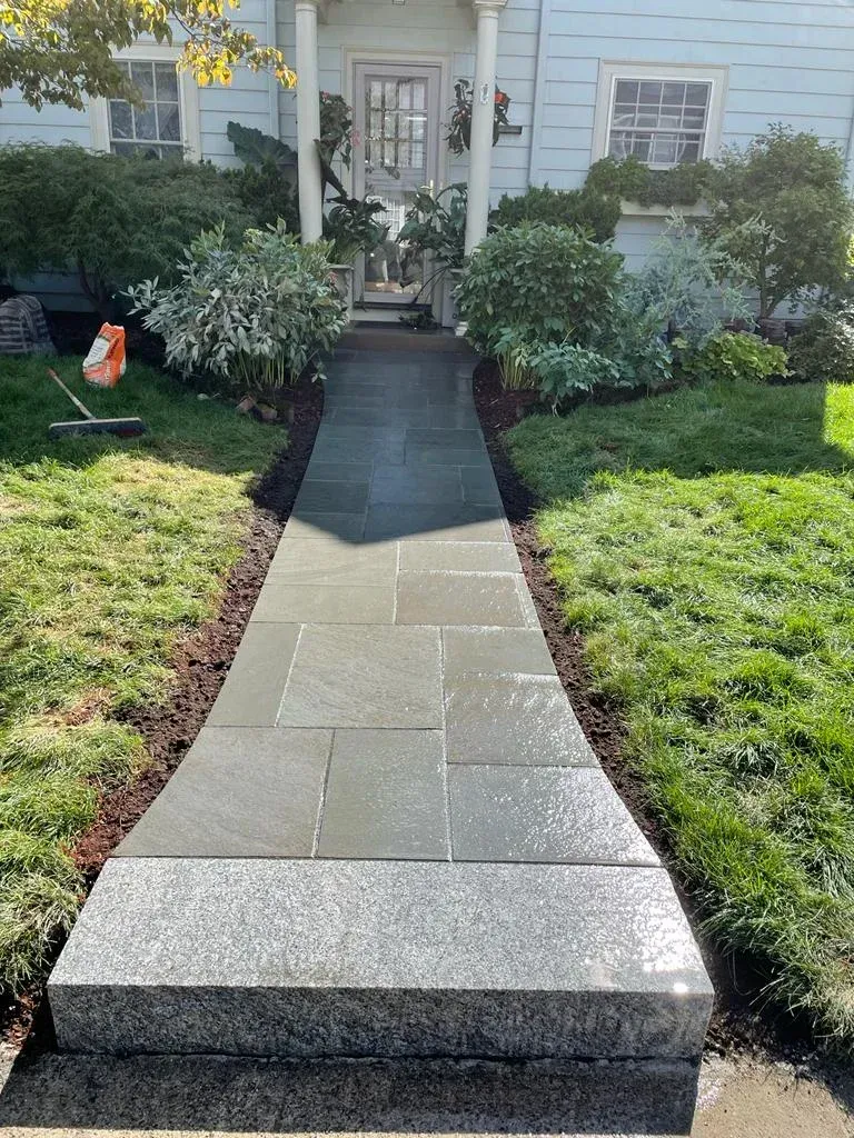 A stone walkway with a wide granite step at the foreground leads to the front door of a white house with landscaping.