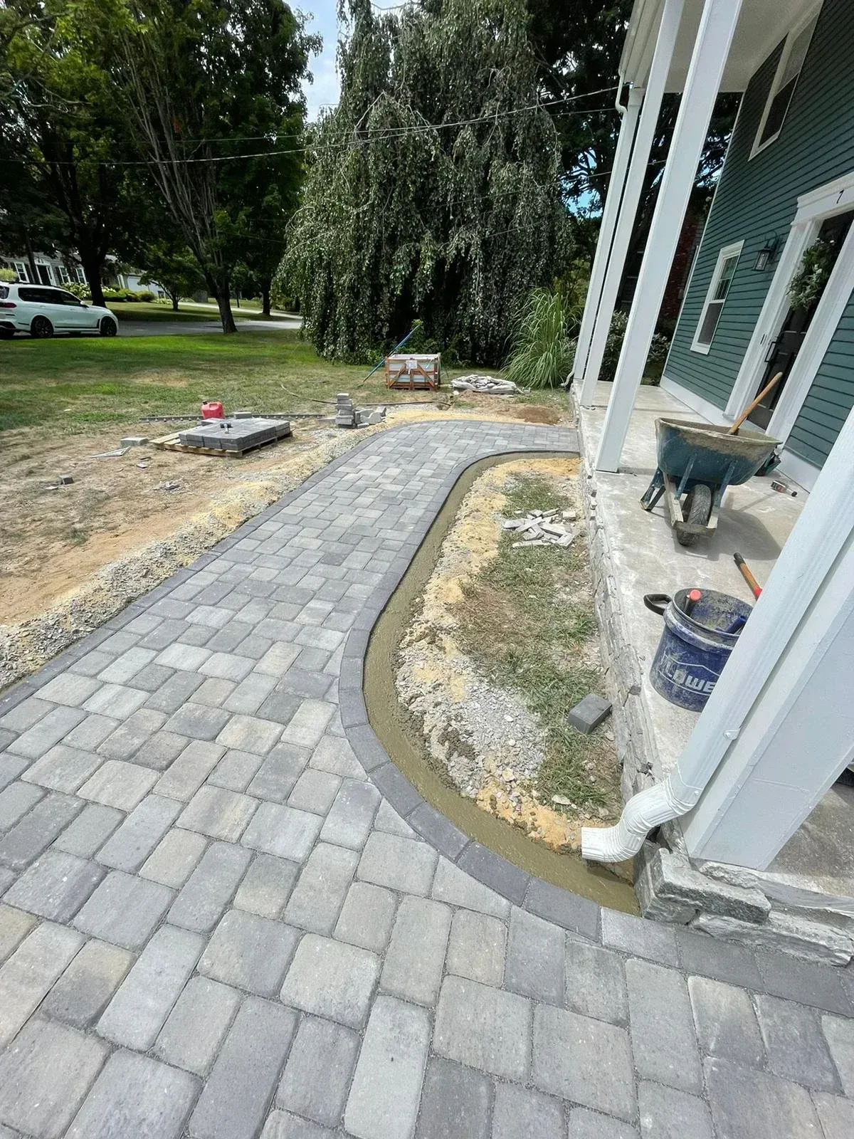 A new grey paver walkway curves toward the porch of a dark green house, with an unpaved border and a wheelbarrow nearby.