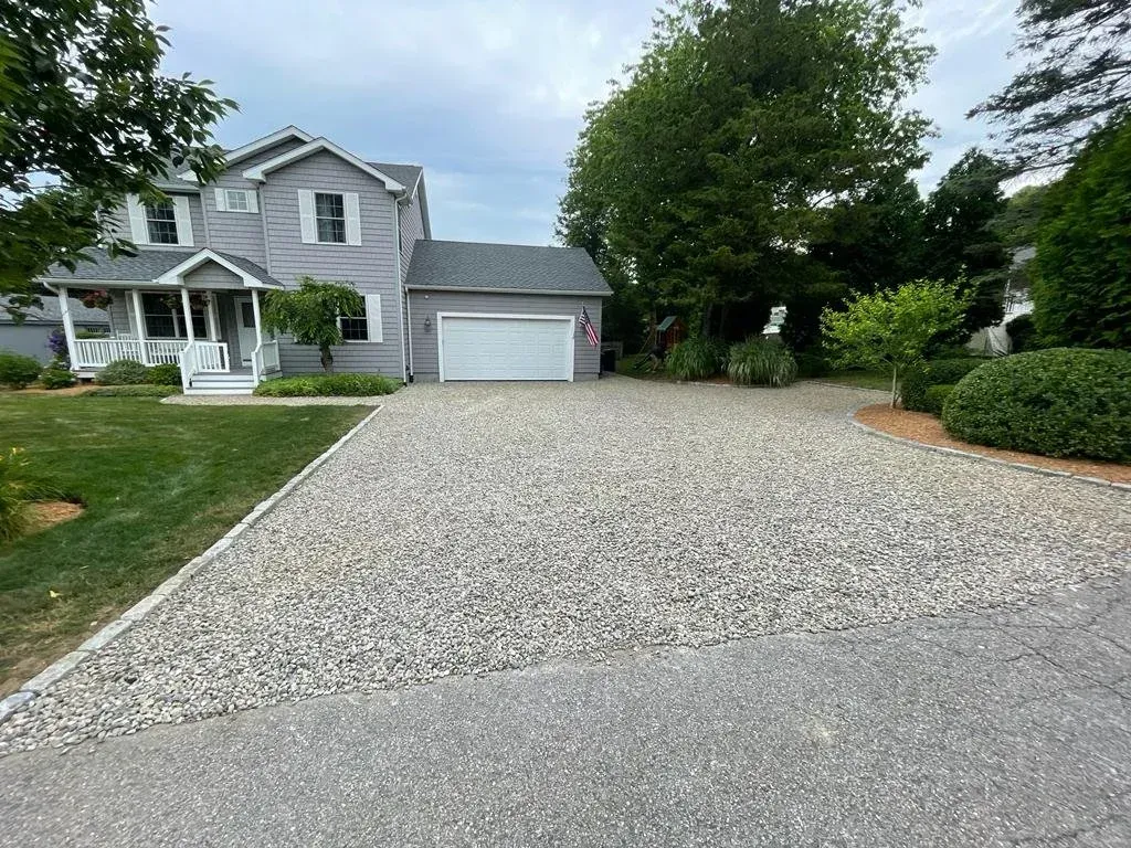 A grey two-story house with a white garage door and a gravel driveway in front of a green lawn and mature trees.
