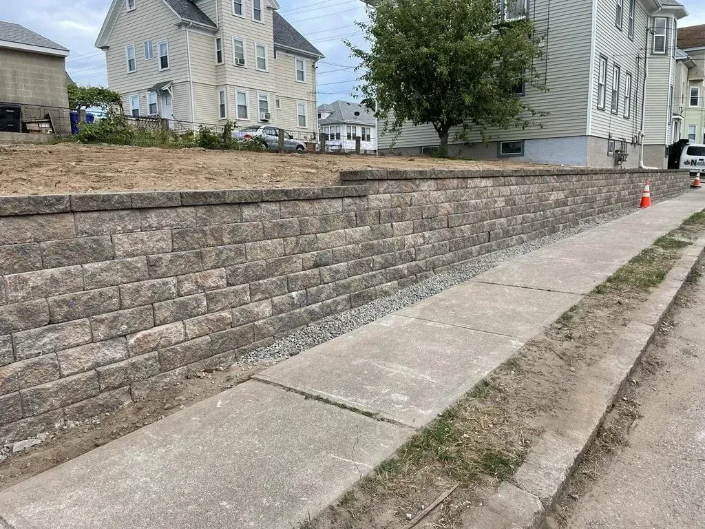 A concrete sidewalk runs along a recently constructed tan stone retaining wall next to residential homes.