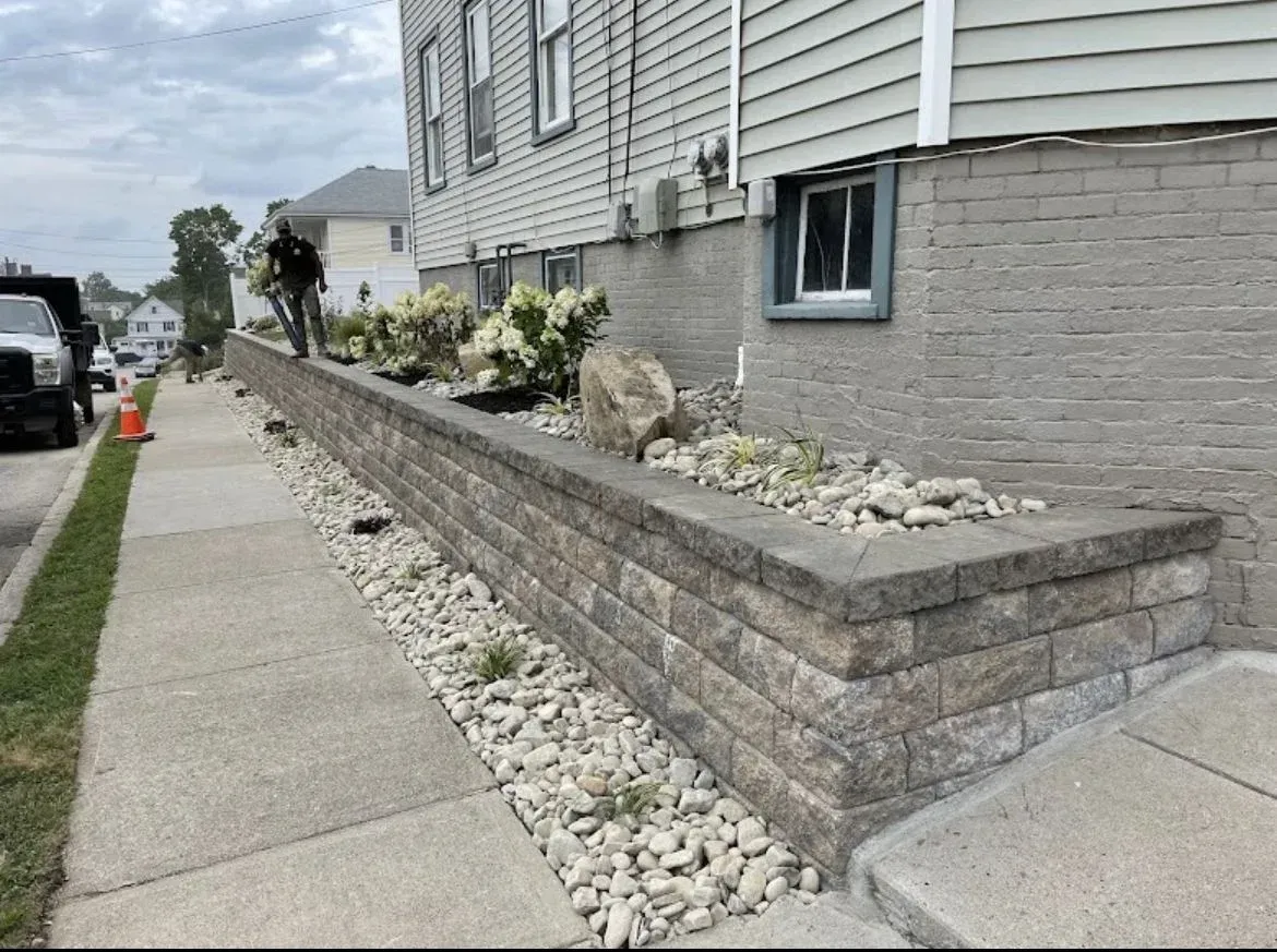 A worker stands near a multi-tiered concrete retaining wall beside a house, bordering a sidewalk filled with stone mulch.