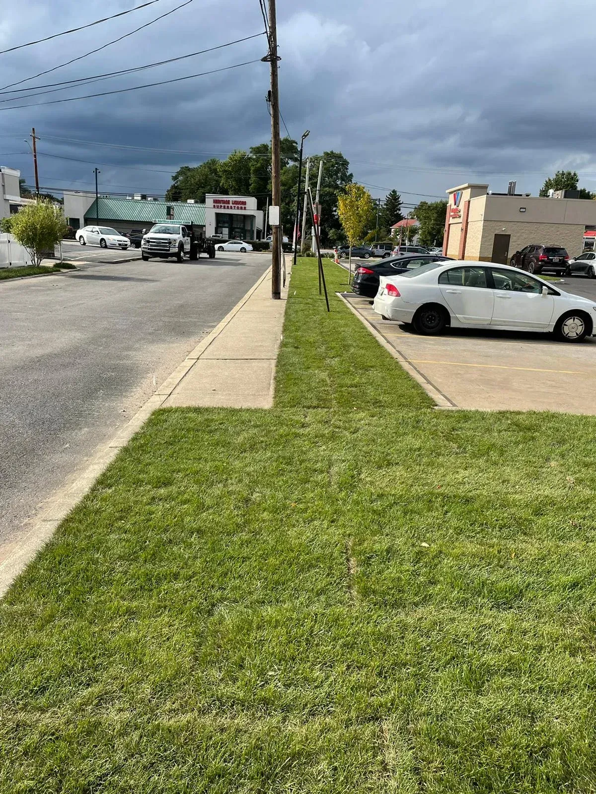A narrow paved sidewalk bordered by grassy areas, separating a street with parked cars from a parking lot under cloudy skies.