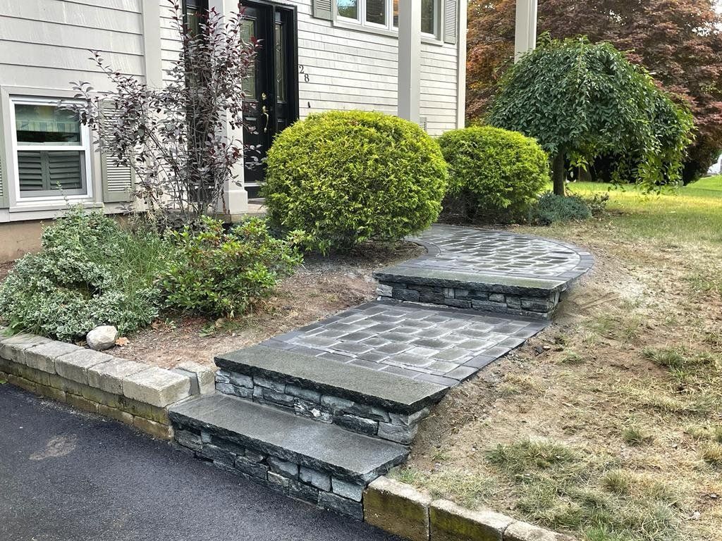 A stone walkway with two steps leads to a house entrance, flanked by green bushes and a retaining wall near a driveway.
