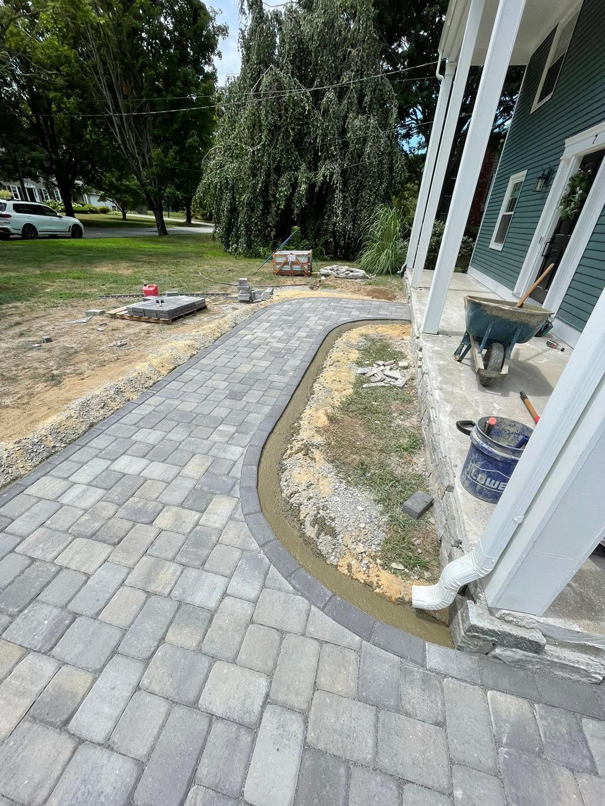 A paved walkway curves alongside a house porch, with an unfinished dirt area and a wheelbarrow nearby.