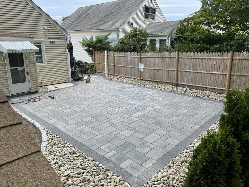 A newly installed gray paver patio bordered by a rock bed and a wooden fence in a suburban backyard.